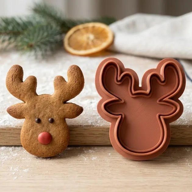 Cortador de galletas con forma de cabeza de reno en tono caramelo junto a una galleta de jengibre, sobre tabla de madera.