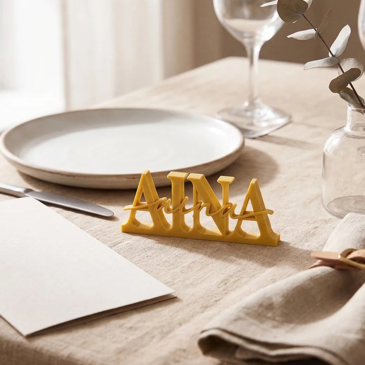 Marcasitios de boda personalizado con el nombre Aina en amarillo mostaza sobre un mantel de lino junto al plato