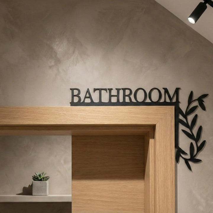 Cartel para esquina de puerta con palabra Bathroom y diseño de hojas en color negro sobre marco de madera.