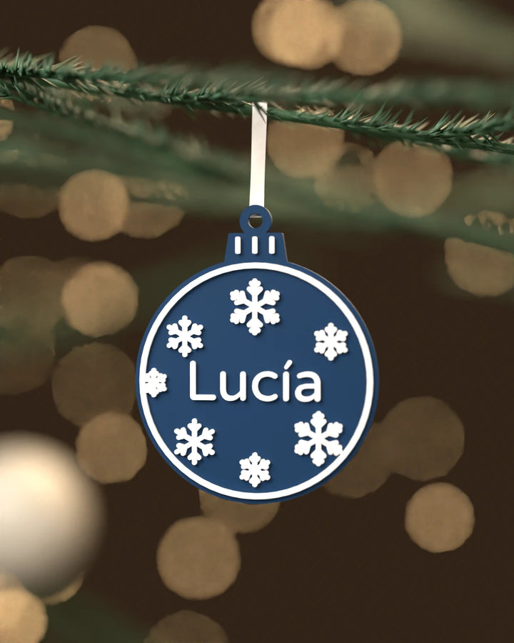 Bola de Navidad Copos con el nombre Lucía en blanco sobre azul marino, colgada de una rama de abeto con luces cálidas