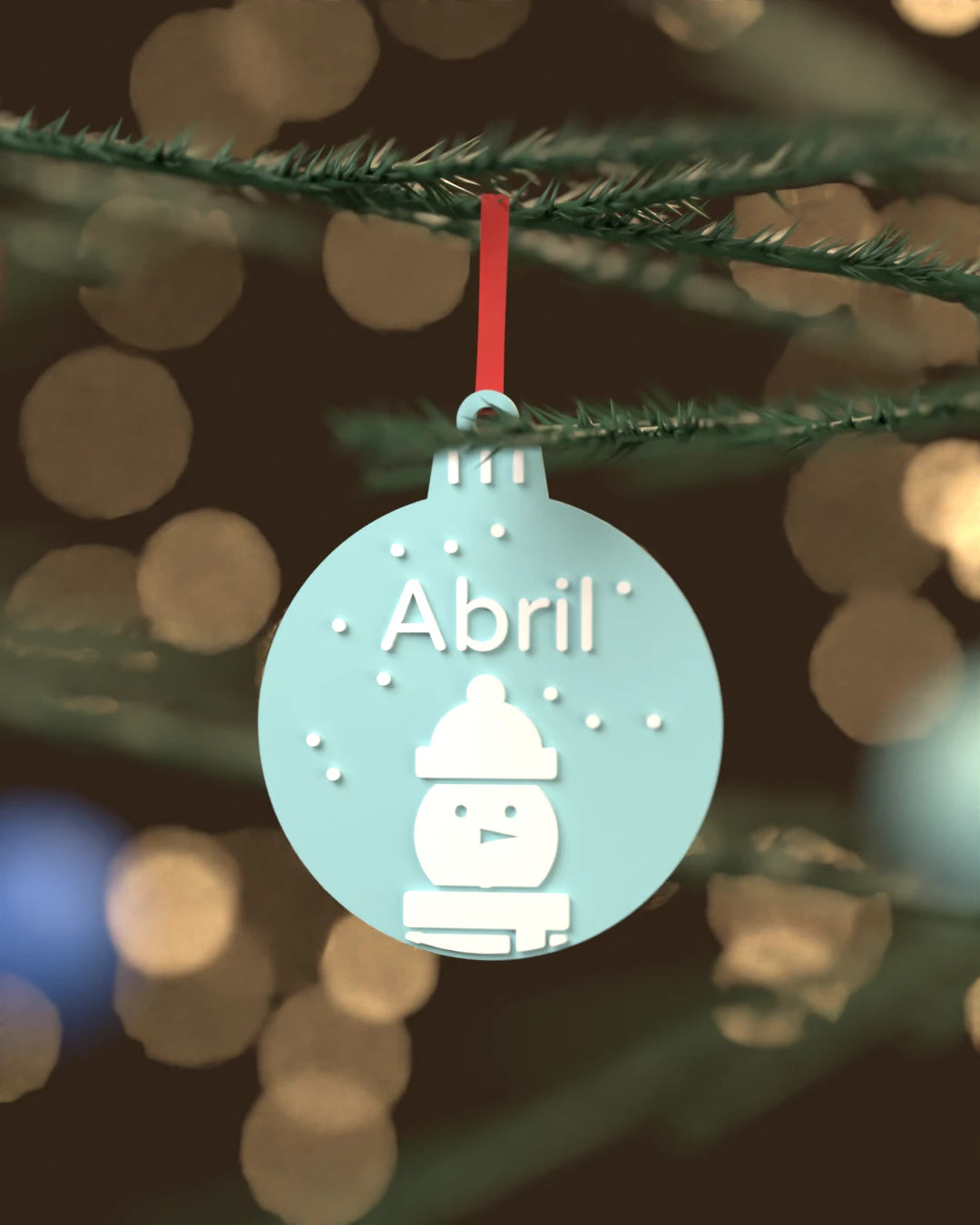 Bola de Navidad Muñeco de Nieve azul hielo con el nombre Abril en blanco, colgada de una rama de abeto
