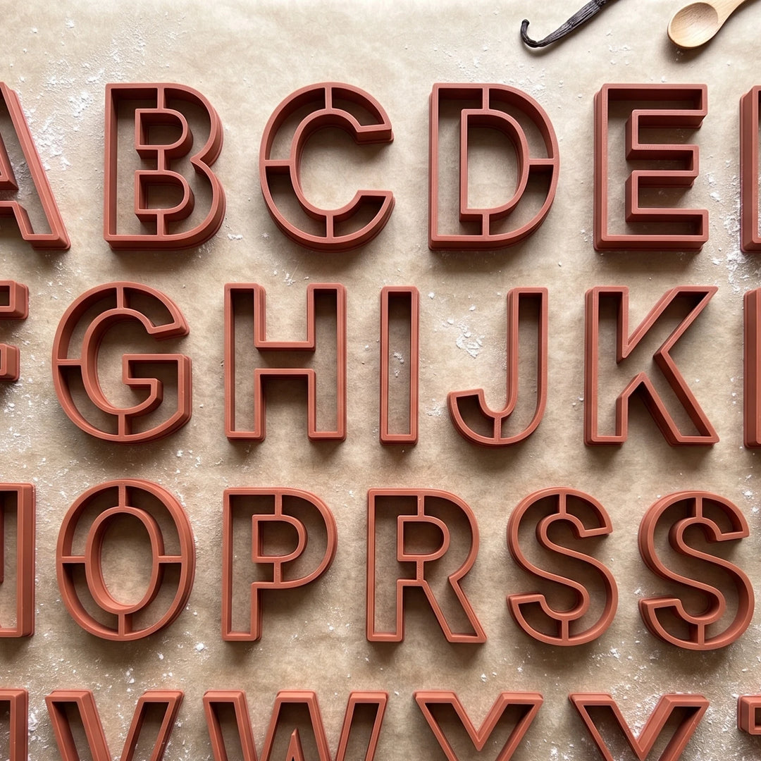 Cortadores de galletas con forma de letras del alfabeto en color terracota sobre papel de horno.