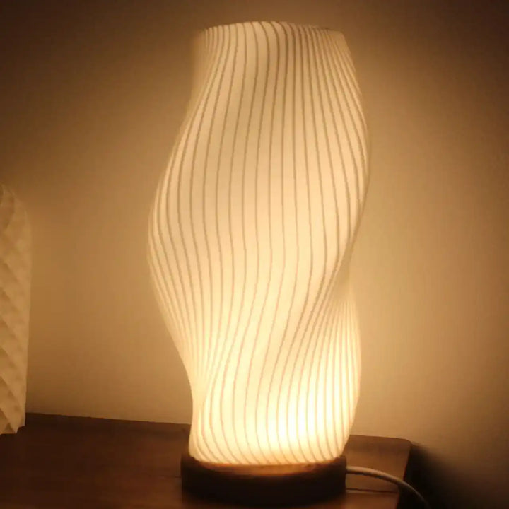 Lámpara de mesa moderna con forma ondulada y textura acanalada blanca, encendida con luz cálida y base redonda.