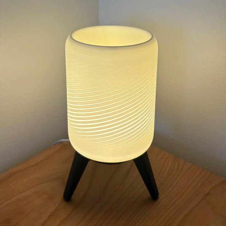 Lámpara de mesa con base trípode y pantalla cilíndrica de efecto espiral. Luz cálida difusa ideal para dormitorios y salones. Diseño contemporáneo y funcional.