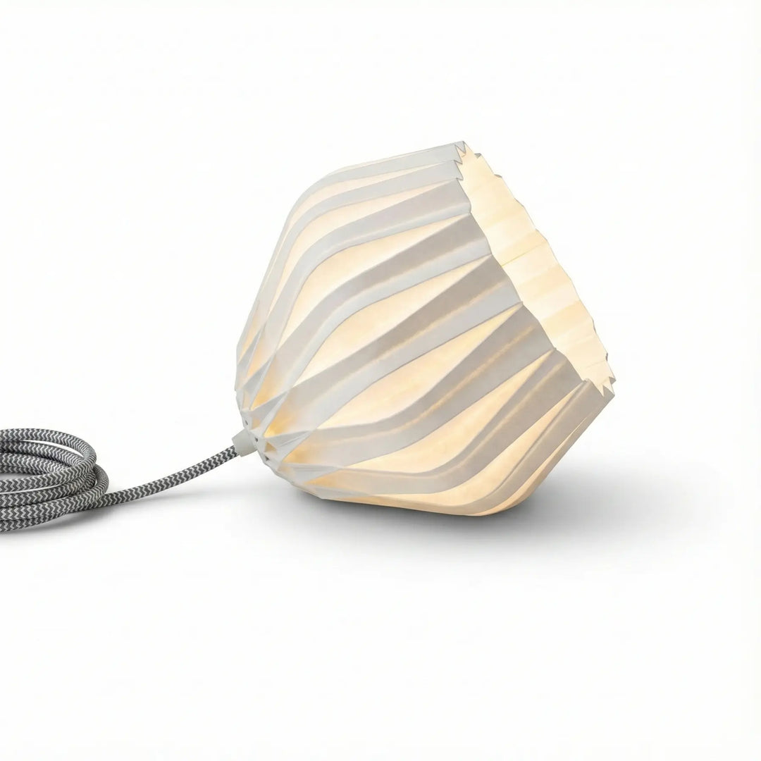 Lámpara de mesa moderna con pantalla blanca facetada y cable textil gris encendida sobre mesita de noche.