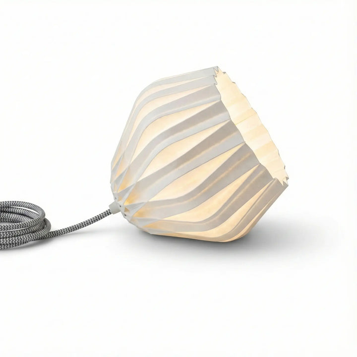 Lámpara de mesa moderna con pantalla blanca facetada y cable textil gris encendida sobre mesita de noche.