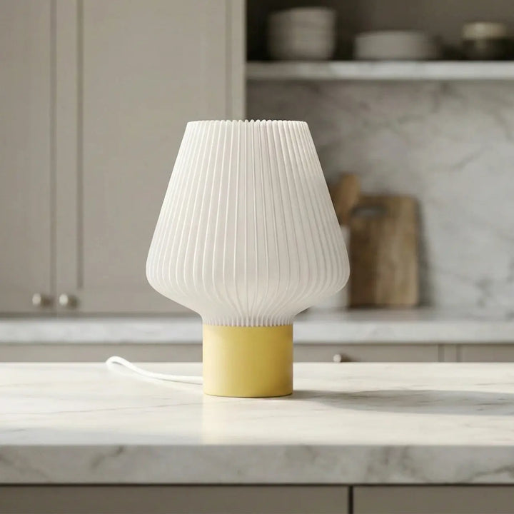 Lámpara de mesa moderna con pantalla plisada blanca y base cilíndrica amarilla sobre encimera de cocina.