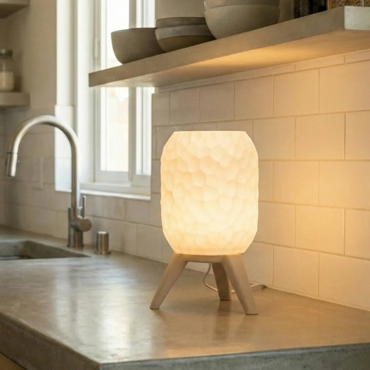 Lámpara de mesa pequeña con pantalla blanca texturizada y patas trípode acabado efecto madera encendida en cocina moderna.