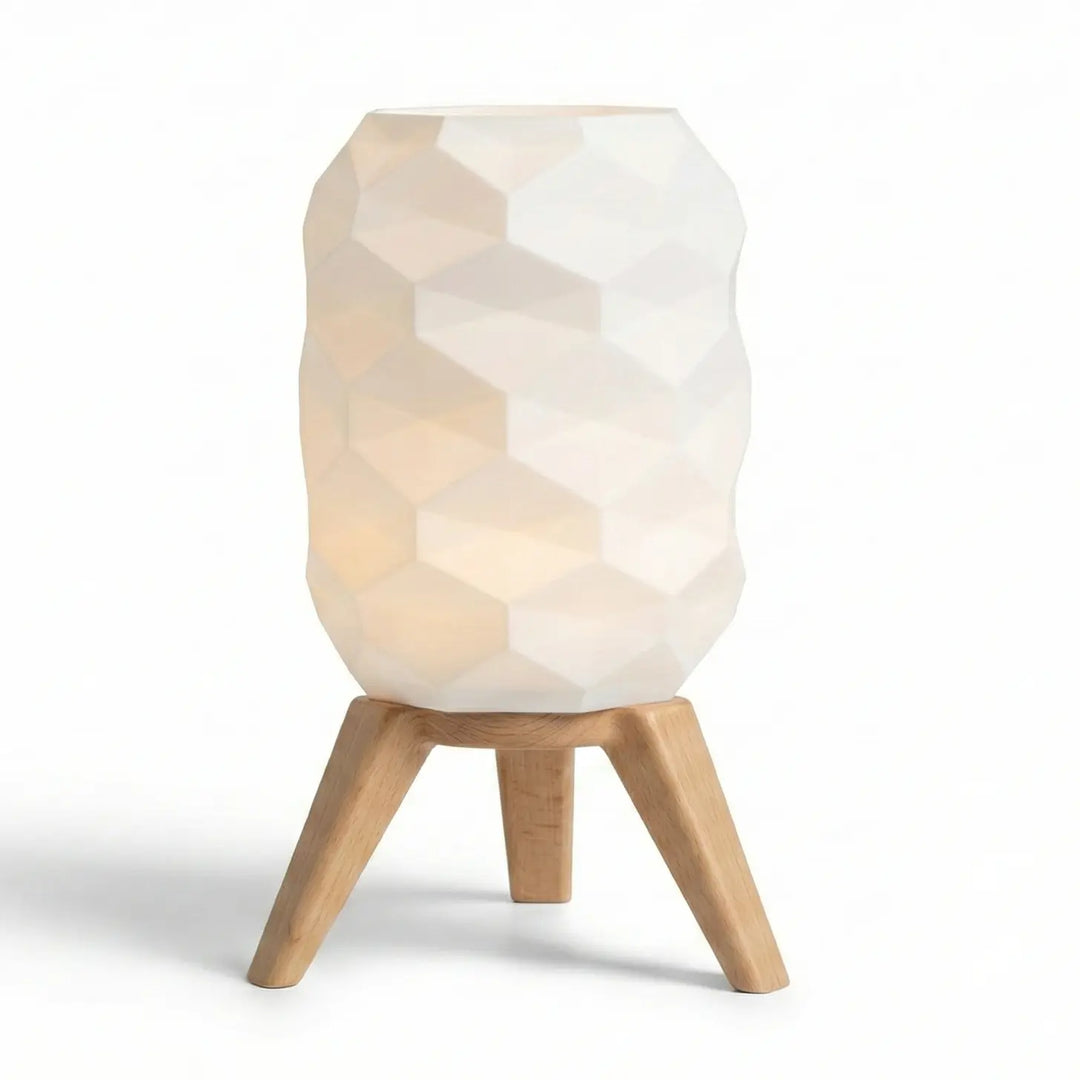 Lámpara de mesa con pantalla geométrica blanca facetada y patas trípode acabado efecto madera sobre mesa auxiliar.