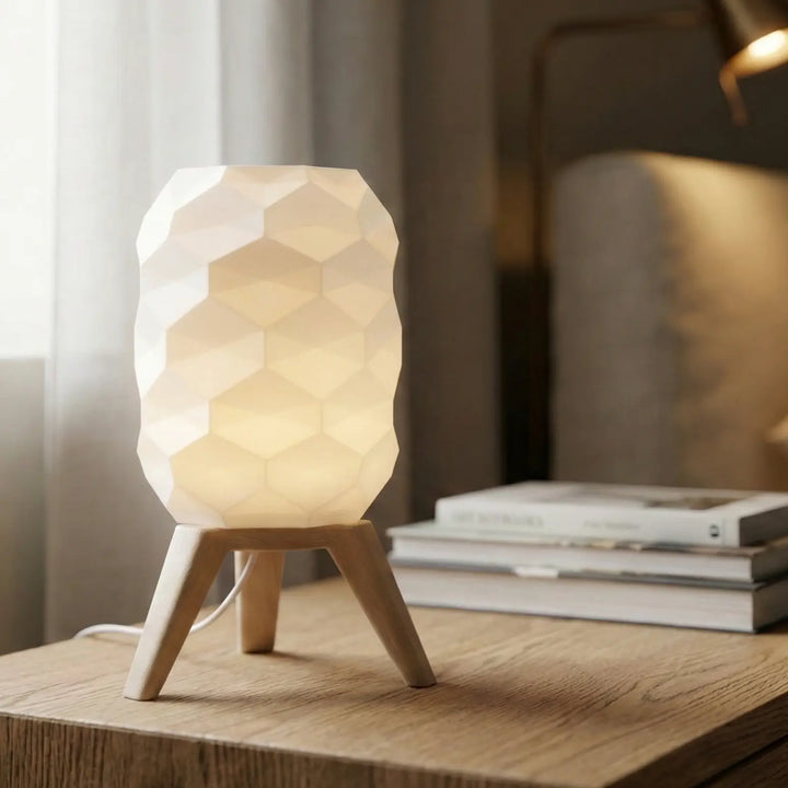 Lámpara de mesa con pantalla geométrica blanca facetada y patas trípode acabado efecto madera sobre mesa auxiliar.