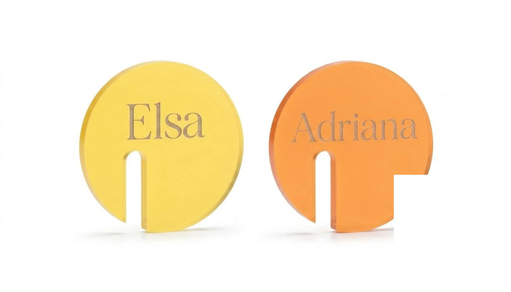 Marcasitios de copa redondos con nombres grabados en tipografía serif sobre fondo blanco en amarillo y naranja