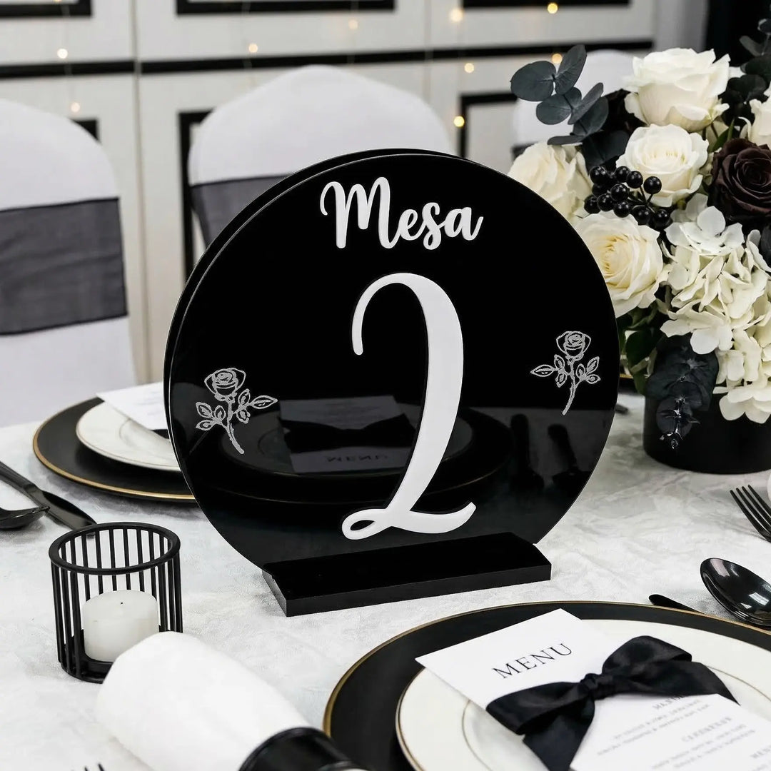 Mesero circular negro brillante con texto en relieve blanco Mesa 2 en mesa de banquete con flores y velas.